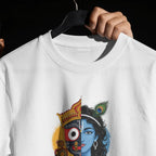 A white crewneck t-shirt on a black hanger, held by a hand against a black background. The shirt features a graphic print of a split deity design, with one half showing a red and gold crown and the other half showing a blue-skinned figure with peacock feather hair ornaments.