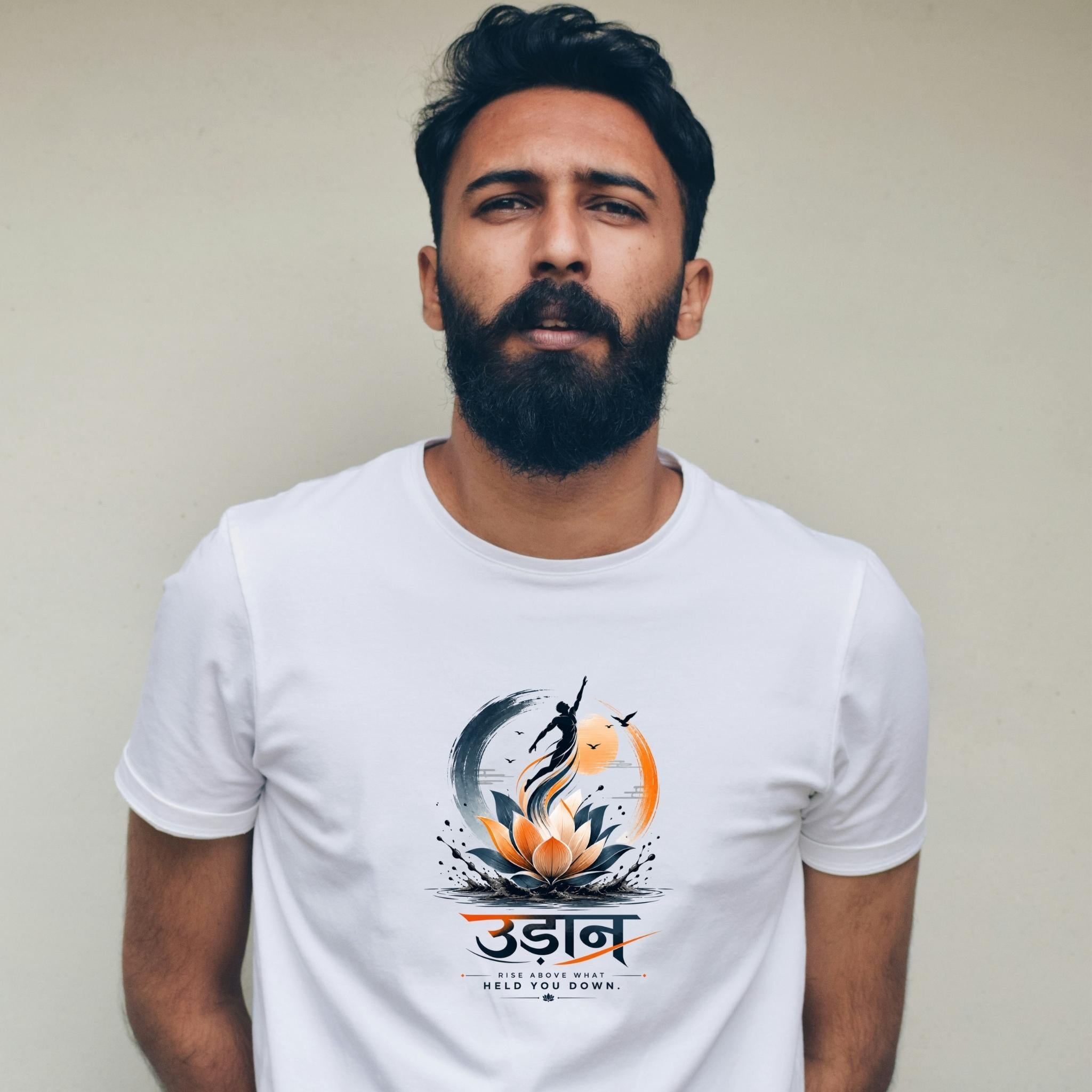 A man with a beard wearing a white crew-neck t-shirt with a colorful graphic print featuring a lotus flower, a fish, and stylized text, set against a plain beige wall.