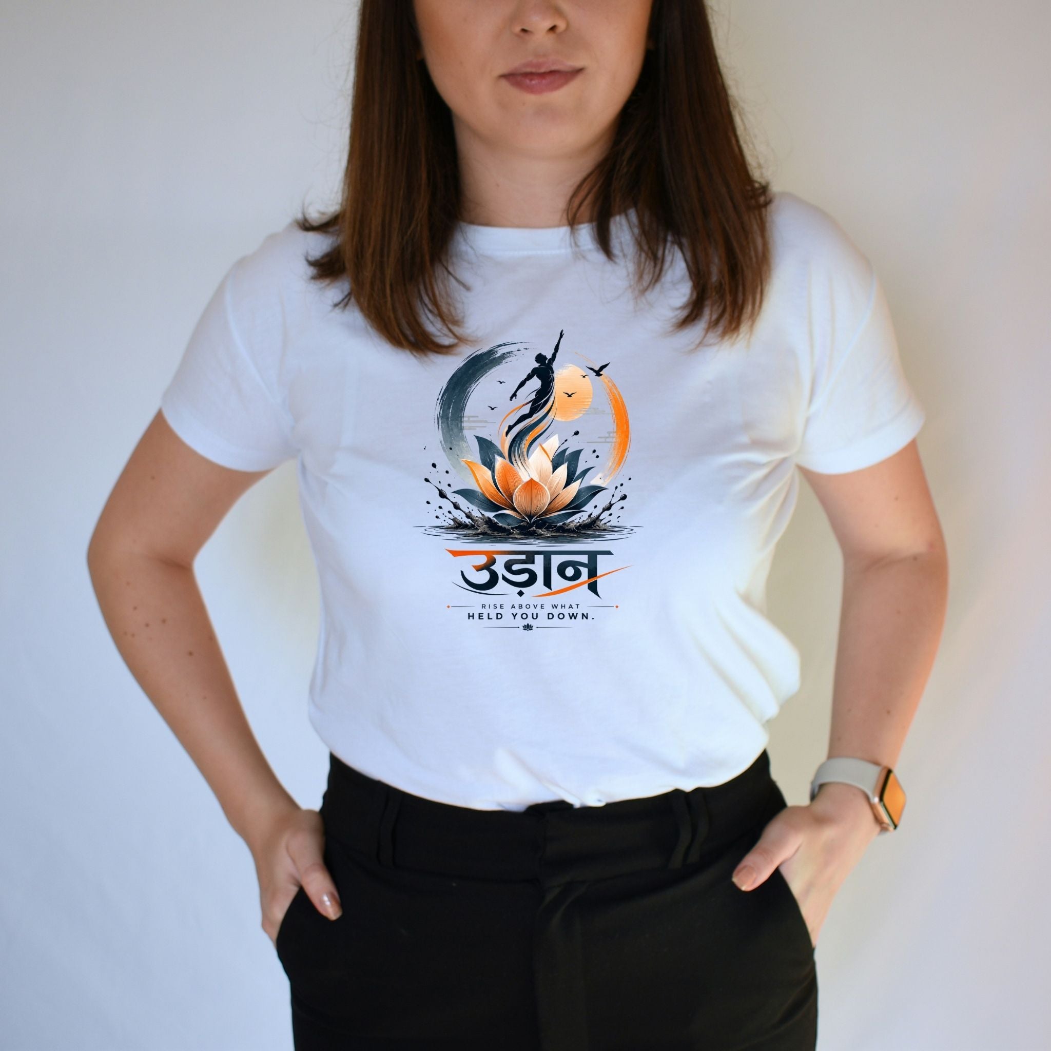 A woman wearing a white short-sleeve graphic T-shirt with a stylized lotus and fish design and the word 'Udaan' printed on the chest, paired with black pants against a plain light background.